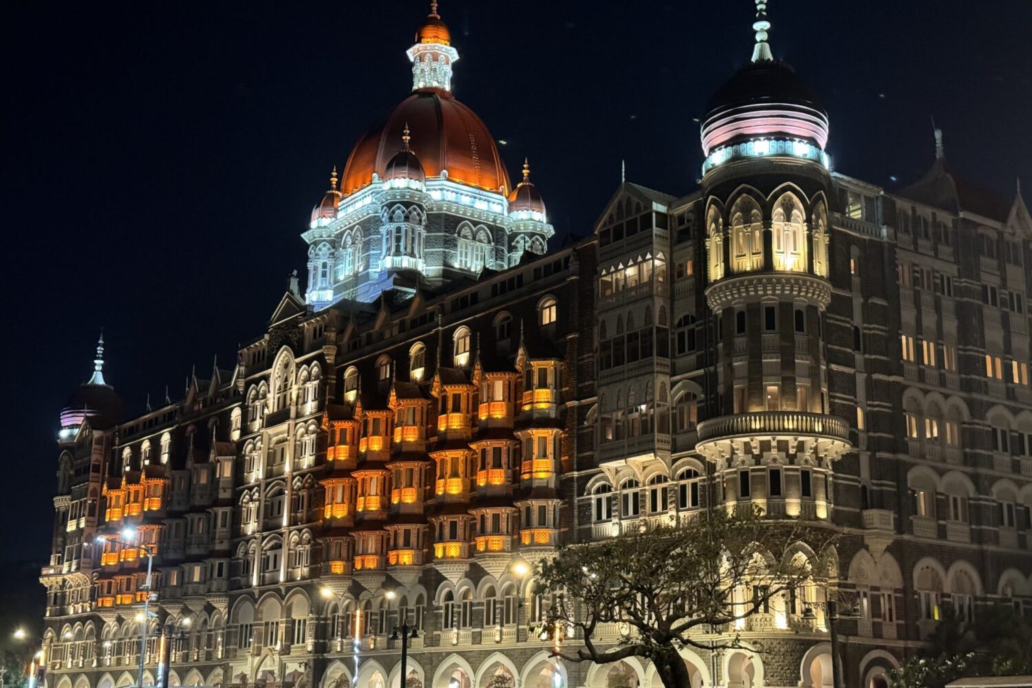 A nighttime view of Taj Mahal Palace hotel in Mumbai, an ornately designed, multi-story building with illuminated domes and arched windows. The building is lit up with warm orange and white lights, highlighting its intricate architectural details.
