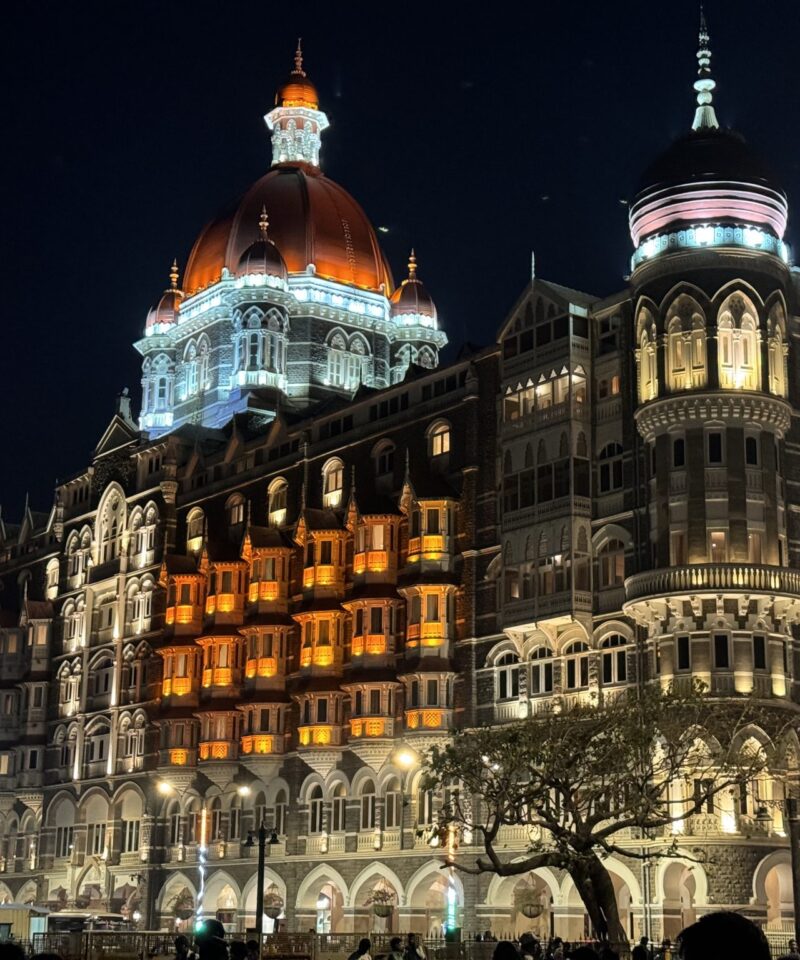 A nighttime view of Taj Mahal Palace hotel in Mumbai, an ornately designed, multi-story building with illuminated domes and arched windows. The building is lit up with warm orange and white lights, highlighting its intricate architectural details.